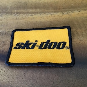Peut inclure: Écusson rectangulaire jaune avec le texte "ski-doo" en noir, souligné de noir. L'écusson a une bordure noire. L'écusson est probablement destiné à une motoneige ou à des vêtements de sport d'hiver.