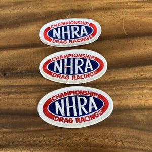 May include: Three embroidered patches with the text "CHAMPIONSHIP NHRA DRAG RACING" in a white oval with red and blue accents. The patches are on a wooden surface.