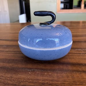 May include: A blue and white speckled ceramic curling stone with a black handle. The stone is round with a flat base and a curved handle on top. The curling stone is on a wooden surface.