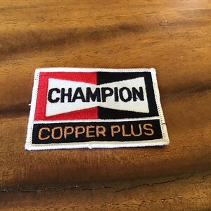 May include: A rectangular patch featuring the word "CHAMPION" in white on a red and black background. Below, "COPPER PLUS" is embroidered in gold. The patch has a white border, and is likely for clothing.