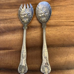 May include: A silver serving spoon and fork set. The spoon and fork heads have a detailed embossed design. The handles feature an ornate, decorative pattern. The set is displayed on a wooden surface.