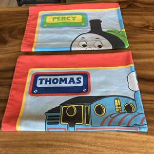 May include: Two pillowcases with red borders and light blue centers. One features Percy the train with the name "PERCY" in a green and yellow box. The other shows Thomas the train with the name "THOMAS" in a blue box.