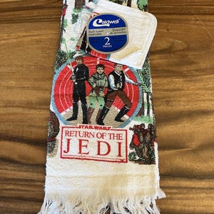 May include: A Star Wars Return of the Jedi bath towel set with a matching washcloth. The towel features a graphic of characters from the movie, with the words "Return of the Jedi" in red. The towel has a white fringe.