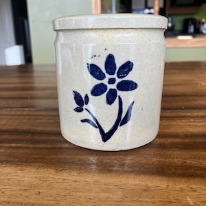 May include: A beige ceramic jar with a lid, decorated with a blue floral motif. The jar has a cylindrical shape and looks handmade. The floral design is a simple, stylized flower with a stem and leaves.