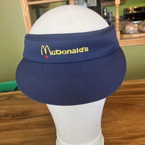 May include: A navy blue visor featuring the McDonald's logo in yellow and red. The visor is displayed on a white mannequin head. The text "McDonald's" is embroidered across the front of the visor.