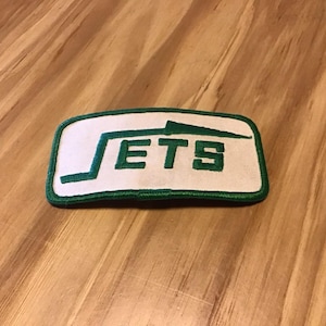 May include: Rectangular patch with the word "JETS" in green, outlined in green, on a white background. The patch has a green border. The patch is on a wooden surface.