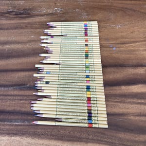 May include: A collection of coloured pencils arranged on a wooden surface. The pencils are beige with colourful stripes and text. The tips of the pencils are various colours, including pink, purple, blue, green, yellow, and red.