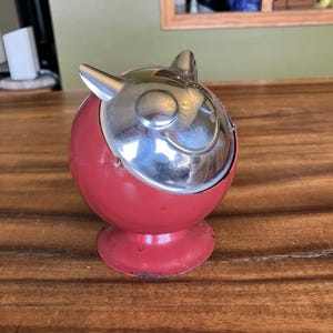 May include: A vintage red ashtray with a chrome lid. The ashtray is round with a small base and two small horns on the top. The chrome lid has a smiling face design. The ashtray is sitting on a wooden surface.