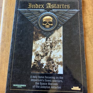 Peut inclure: Un livre noir intitulé "Index Astartes" avec un emblème de crâne doré. La couverture présente une illustration de Space Marines et le texte "A holy tome focusing on the Imperium's finest warriors, the Space Marines of the Adeptus Astartes."