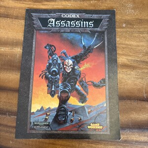 Peut inclure: Un supplément Codex: Assassins de Warhammer 40,000. La couverture présente un guerrier futuriste au visage de crâne, tenant une grosse arme et des lames. L'arrière-plan est une scène orange et rouge flamboyante avec le logo Games Workshop.