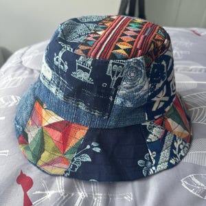 May include: A patchwork bucket hat with a variety of patterns and colors. The hat features a mix of blue denim, geometric shapes, and floral designs. The crown has a colorful woven fabric.