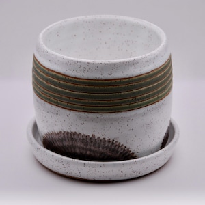 May include: A speckled white ceramic planter with a matching saucer. The planter features horizontal bands of green and brown, and a dark brown, textured design on the saucer. The planter is designed for indoor or outdoor use.