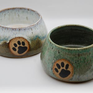 May include: Two ceramic pet bowls. One is light blue and brown, the other is green. Each bowl has a paw print design. The bowls are round with a wide opening and a slightly tapered base.