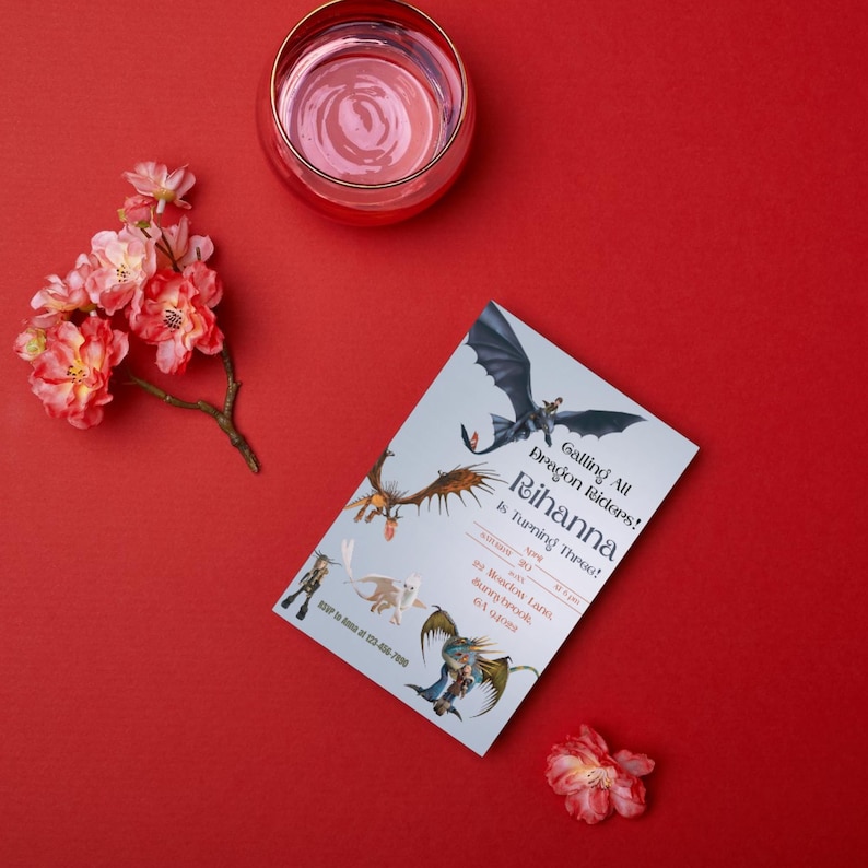 May include: A birthday invitation featuring dragon illustrations and the text "Calling All Dragon Riders! Rihanna Is Turning Three!" is displayed on a red surface. A glass of pink liquid and pink flowers are also present.