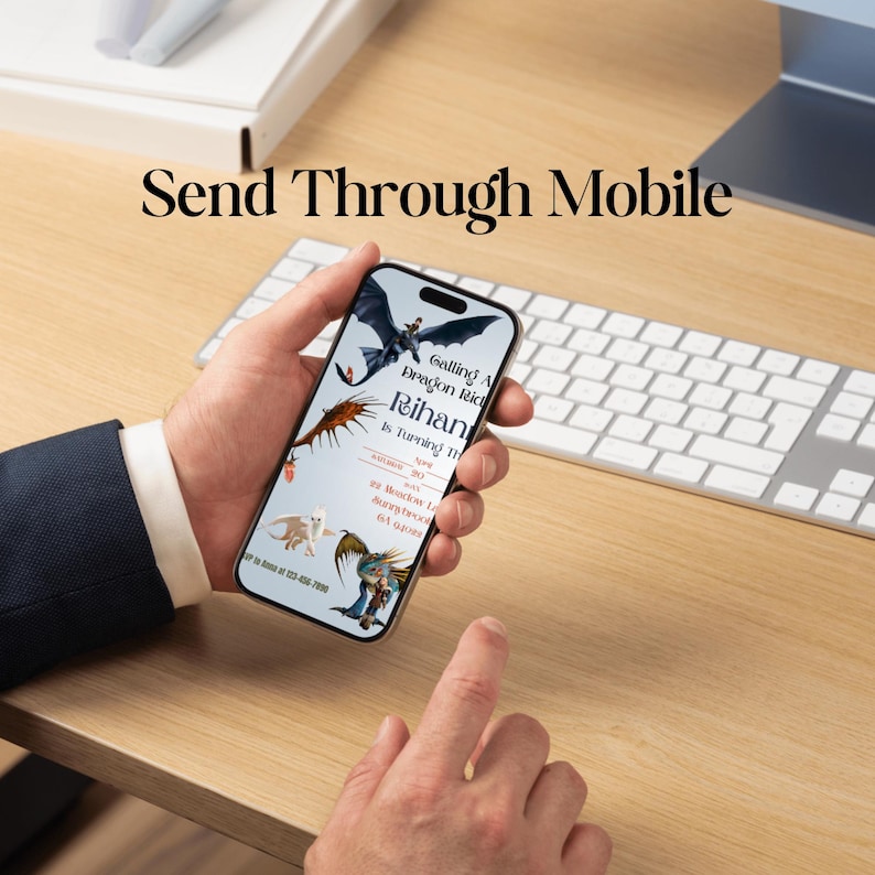 May include: A smartphone screen shows a birthday invitation with dragon graphics. The phone is held in a hand, with a keyboard and wooden desk in the background. The invitation text reads "Calling All Dragon Riders". The text "Send Through Mobile" is at the top.