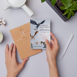 May include: A light blue invitation card featuring dragon illustrations and the text "Calling All Dragon Riders! Rihanna is Turning Three!" held by hands. A brown envelope and a white pen are also visible.