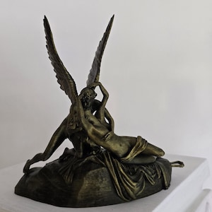 Puede incluir: Una escultura de color bronce que representa una figura alada abrazando a una figura reclinada. La escultura presenta alas detalladas y tela drapeada, sobre una base texturizada. Las figuras están en un abrazo.