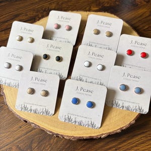 May include: A collection of ten pairs of round stud earrings displayed on individual white cards labeled J. Pease Collections. The earrings feature various solid colors including pink, gold, white, black, red, blue, and beige, each with a metallic rim.
