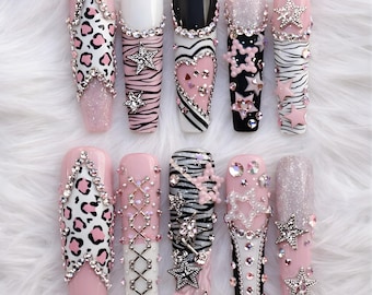 Op de afbeelding: Een set van tien lange, kunstmatige nagels met diverse ontwerpen. De nagels hebben roze, witte en zwarte bases, versierd met glitter, strass en stervormige versieringen. Ontwerpen omvatten luipaardprint, zebra strepen en hartpatronen.
