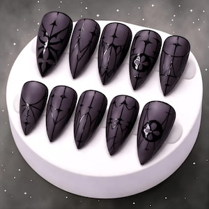 May include: A set of ten matte gray press-on nails with black gothic-style designs. Each nail features a different pattern, including geometric shapes and floral motifs. The nails are almond-shaped and displayed on a white circular platform.