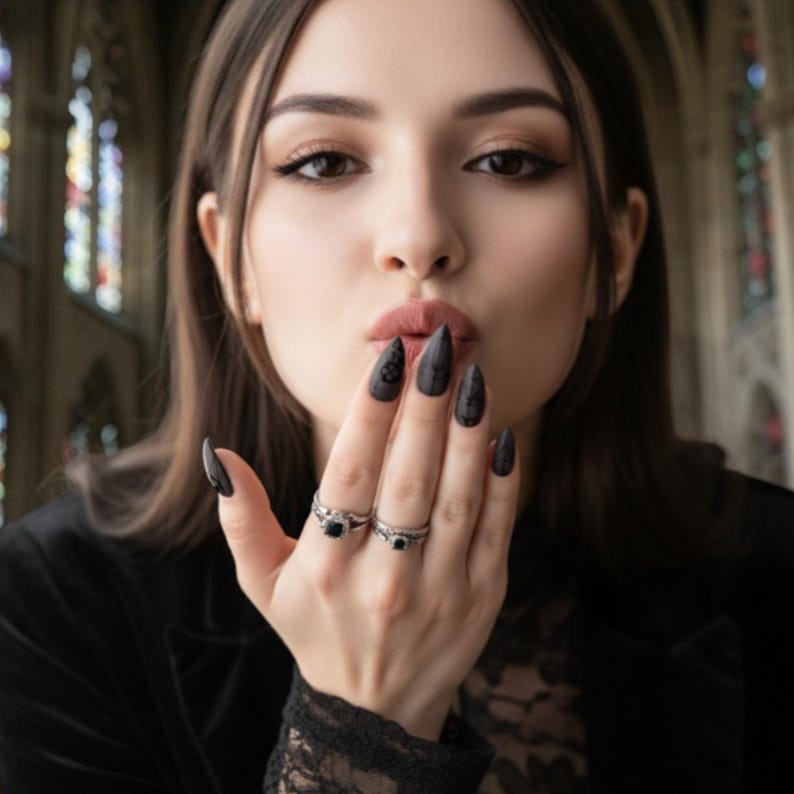 May include: Black stiletto nails with intricate designs. Two silver rings with black gemstones. The image features a woman blowing a kiss, with a black jacket and lace top. The background includes stained glass windows.