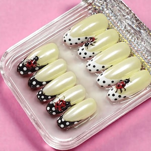 May include: Artificial nails with a pale yellow base and a black and white polka dot design on the tips. Each nail features a red ladybird design. The nails are arranged in a clear, rectangular container.