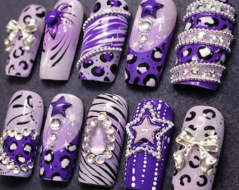 May include: A collection of ten artificial nails in various designs. The nails feature shades of purple, white, and silver, with embellishments like bows, stars, rhinestones, and animal prints. The designs include zebra stripes, leopard spots, and abstract patterns.