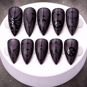 May include: A set of ten matte black, almond-shaped false nails. Each nail features a unique black geometric design, including floral and linear patterns. The nails are arranged on a white, circular surface.
