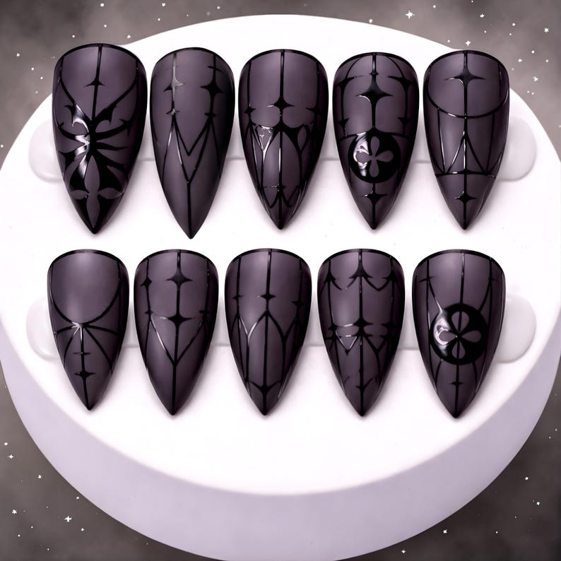 May include: A set of ten matte black, almond-shaped false nails. Each nail features a unique black geometric design, including floral and linear patterns. The nails are arranged on a white, circular surface.