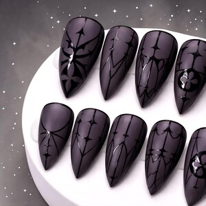 May include: A set of ten matte gray artificial nails with black gothic-style designs. Each nail features a different pattern, including floral and geometric shapes. The nails are almond-shaped and arranged on a white surface.