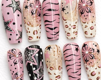 May include: A collection of ten artificial nails with various designs. The nails feature pink, black, and glitter accents, including stars, floral patterns, and animal prints. Each nail is a different design.