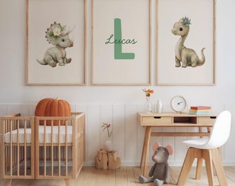 May include: Three framed art prints featuring watercolor dinosaur illustrations. One print shows a triceratops, another a green letter "L" with the name "Lucas", and the third a brontosaurus. The prints are displayed above a wooden crib and desk.