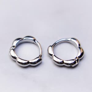 May include: Two silver-colored hoop earrings with a unique, rounded design. Each earring features a series of smooth, petal-like shapes around the hoop. The earrings are displayed on a white marble surface, creating a clean and elegant presentation.