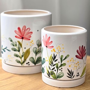 May include: Two cylindrical white ceramic planters of different sizes, each featuring a painted botanical design with pink flowers, green foliage, and small yellow accents. Both planters include a matching circular base tray for drainage.