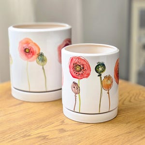 Op de afbeelding: Twee cilindrische plantenpotten van wit keramiek met bijpassende schotels, voorzien van aquarelillustraties van rode klaprozen en groene zaaddozen. De potten zijn glad, glanzend en hebben een open bovenkant voor kamerplanten.