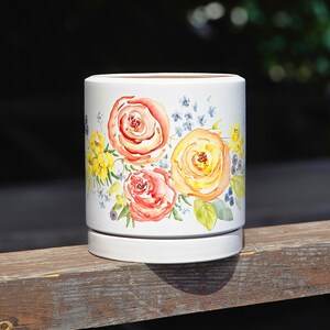 May include: A white cylindrical ceramic planter featuring a watercolour floral design. The artwork displays large orange and yellow roses surrounded by smaller blue flowers, green leaves, and dark berries. The planter includes a matching circular base.