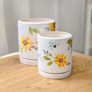 Può includere: Due vasi cilindrici in ceramica bianca con sottovasi abbinati, decorati con motivi di girasoli gialli dipinti a mano, foglie verdi e piccoli accenti floreali blu. I vasi presentano una finitura liscia e lucida.