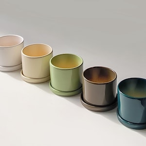 May include: Five ceramic planters in a row, each with a matching saucer. The planters are cylindrical with a glossy finish, and come in a variety of colors: white, cream, green, brown, and teal. Great for small plants.