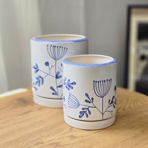 May include: Two cylindrical ceramic planters with a white base and blue hand-painted botanical illustrations of flowers and leaves. Each planter features a matching blue rim and a circular saucer base, showcasing a clean and simple design.