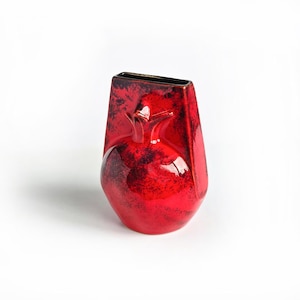 May include: A ceramic vase with a vibrant red glaze and dark, mottled black accents. The vessel features a distinct pomegranate-shaped relief on the front, a rectangular top opening, and a smooth, glossy finish across its entire surface.