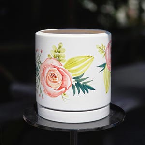 Könnte beinhalten: Ein zylindrischer weißer Keramikübertopf mit handgemaltem Blumenmuster. Das Design zeigt große rosa Rosen, gelbe Knospen und grünes Blattwerk, die das Äußere verzieren. Der Übertopf wird mit einem passenden runden Untersetzer geliefert.