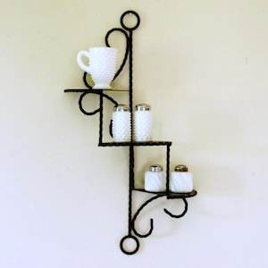 May include: A black wrought iron wall shelf with a decorative design, holding a white milk glass creamer and salt and pepper shakers. The shelf has a spiral and loop design. The milk glass items have a hobnail pattern.