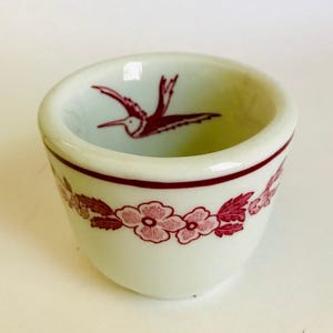 May include: A small, off-white ceramic cup with a burgundy floral and bird design. The cup features a burgundy band around the rim and a floral pattern around the base. A burgundy bird is depicted inside the cup.