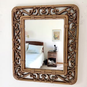 May include: Square decorative mirror with a brown, ornate, woven frame. The frame features intricate scrollwork and a rectangular shape. The mirror is hanging on a white wall, reflecting a bedroom scene.