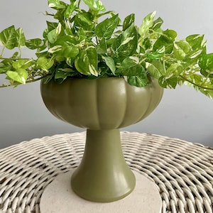 Puede incluir: Una jardinera verde oliva con un cuenco en forma de calabaza, llena de vides de hojas verdes y blancas. La jardinera se asienta sobre una base redonda blanca. El fondo es gris claro.