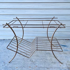 May include: A vintage, two-tiered metal plant stand with a gold finish. The stand features a rectangular top and a lower shelf with a zig-zag design. The curved legs and decorative ball accents add to its mid-century modern aesthetic.