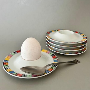 May include: A white egg in a white egg cup with a colorful geometric border, a silver spoon, and a stack of matching egg cups. The egg cups have a vibrant design with red, yellow, blue, and green accents.