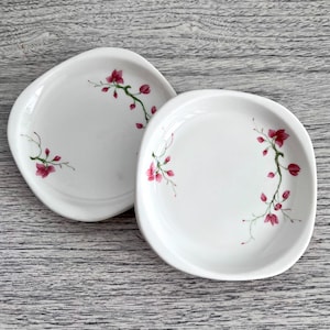 May include: Two white ceramic plates with a floral design. Each plate has a unique, irregular shape and is decorated with a delicate pink flower and green stem pattern. The plates are set against a light gray wooden surface.