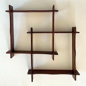 May include: A dark brown wooden wall shelf featuring an interlocking grid design with three horizontal shelves supported by three vertical panels. The vertical panels have curved, wavy edges, creating a geometric structure with multiple open compartments.