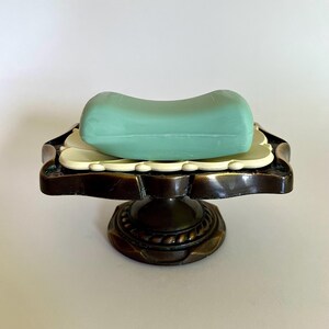 May include: A rectangular, light green bar of soap rests on a cream-colored, scalloped-edge tray. This tray is supported by a dark, antique bronze-finished metal pedestal base featuring a circular, rope-textured design around the central stem.